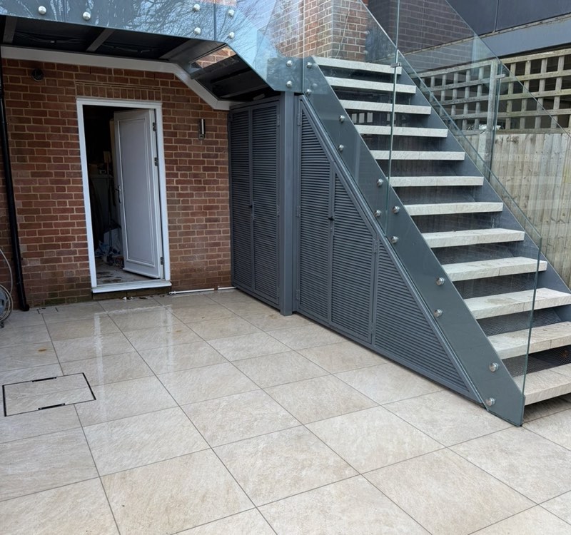 Grey steel staircase with stone treads and a frameless glass balustrade rising from a tiled courtyard