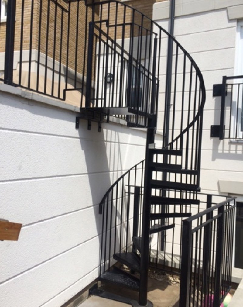 Black spiral staircase with vertical bar balustrade rising from a courtyard to a first-floor terrace